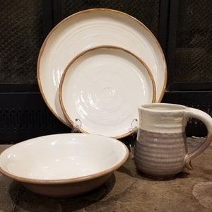 May include: A cream-colored ceramic dinnerware set with a rustic design. The set includes a large plate, a smaller plate, a bowl, and a mug. Each piece has a brown rim. The mug has a two-tone design with a ribbed lower section.