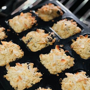 May include: A black muffin tin filled with golden brown muffins topped with shredded coconut. The muffins are arranged in a grid pattern.