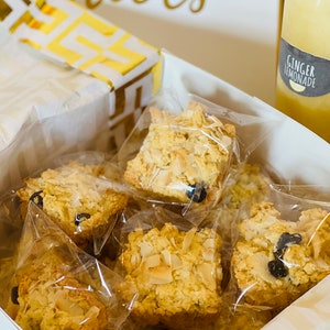 May include: A white gift box filled with individually packaged coconut and raisin scones. The box is decorated with a gold and white patterned paper. The box is sitting on a white surface next to a bottle of ginger lemonade.