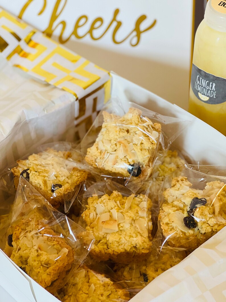 May include: A white gift box filled with individually packaged coconut and raisin scones. The box is decorated with a gold and white patterned paper. The box is sitting on a white surface next to a bottle of ginger lemonade.