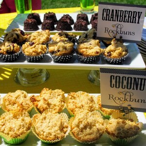 May include: A white platter with two rows of small, round, baked goods. The top row has cranberry rock buns and the bottom row has coconut rock buns. Each row has a black and white sign with the flavor and "Rockbuns" written on it.