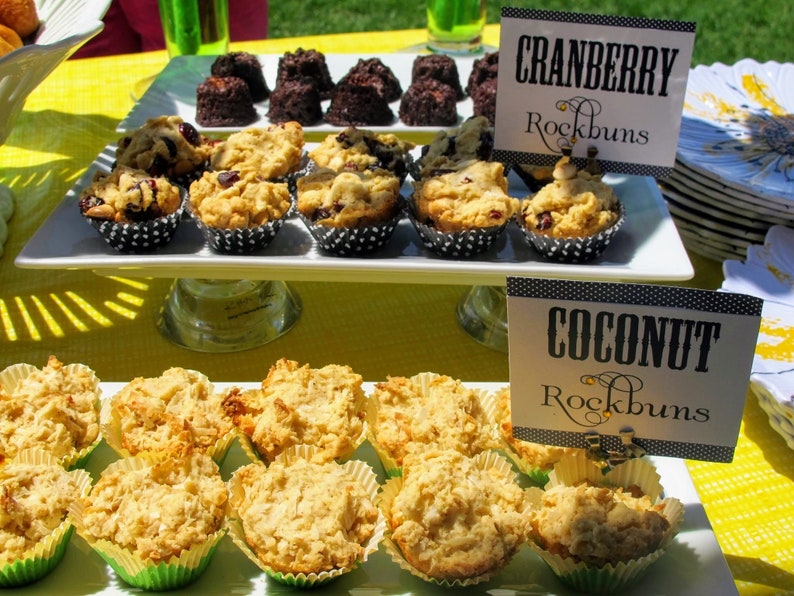 May include: A white platter with two rows of small, round, baked goods. The top row has cranberry rock buns and the bottom row has coconut rock buns. Each row has a black and white sign with the flavor and "Rockbuns" written on it.