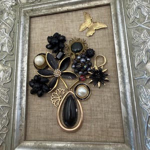 May include: A framed wall art piece featuring a collection of vintage jewelry arranged to resemble a floral bouquet. The composition includes black beads, faux pearls, gold-tone metal accents, and a gold butterfly brooch, all set against a beige fabric background.
