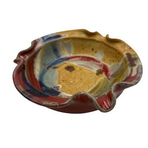 Puede incluir: Un cuenco de cerámica hecho a mano con un borde ondulado. El cuenco presenta una vibrante paleta de colores de rojo, azul, amarillo y marrón. El interior tiene pinceladas abstractas y el exterior es de color rojo intenso.