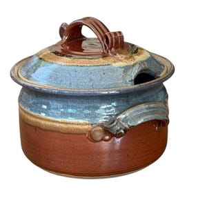 May include: A handmade ceramic jar with a lid. The jar is primarily reddish-brown with a blue and tan band around the middle. The lid has a brown handle and a blue and tan design. The jar has a small opening on the side.