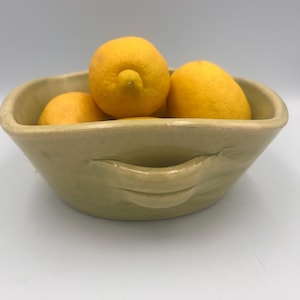 May include: A light green ceramic bowl with a square shape, holding three bright yellow lemons. The bowl has a textured surface and a handle-like design on the side. The lemons are a vibrant contrast to the bowl's color.