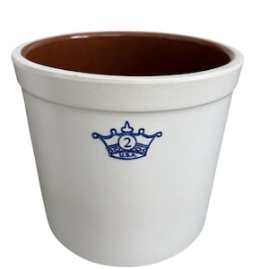 Puede incluir: Olla de cerámica blanquecina con interior marrón. Un emblema de corona azul con el número "2" y "U.S.A." está impreso en la parte delantera. La olla tiene una abertura ancha y una forma ligeramente cónica, adecuada para la cocina o la decoración.