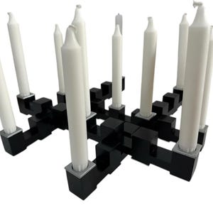 May include: A black geometric candle holder with multiple white candles. The candle holder has a modern design with a series of interconnected black blocks. The white candles are of varying heights.
