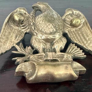 May include: A gold-toned brass eagle wall plaque featuring a spread-wing eagle perched above a blank scroll banner. The eagle has detailed feathers and two small spherical accents on its wings. The metal surface shows a weathered patina.