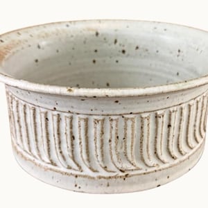 May include: A handmade, off-white ceramic bowl with a wide rim and a textured, vertical ribbed design around the exterior. The bowl has small, dark speckles throughout and a slightly rough, rustic appearance, suggesting a natural, artisanal craft.