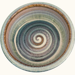 May include: A round ceramic bowl featuring a reactive glaze with a swirling pattern in shades of blue, brown, and cream. The interior surface displays a prominent spiral design radiating from the center, while the rim has a smooth, glossy finish.