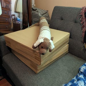 May include: A handmade crochet dachshund dog in brown and white, lying on top of two stacked cardboard boxes. The dog has floppy ears and a long body. The boxes are a light brown color, and the background includes a gray couch and a wooden cabinet.