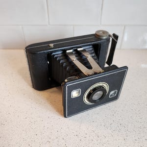 May include: A black vintage folding camera with a silver lens and a silver knob on the top. The camera is on a white countertop.