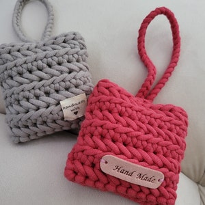 Può includere: Due cuscini a forma di cuore all'uncinetto con anelli per appenderli. Uno è grigio con un'etichetta "fatto a mano con amore" e l'altro è rosso con un'etichetta "fatto a mano".