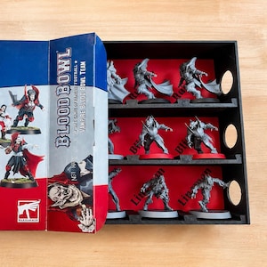 Puede incluir: Una caja de juego de mesa Blood Bowl Vampire Blood Bowl Team con ilustraciones de figuras de vampiros con uniformes rojos y blancos. Junto a la caja hay una bandeja de almacenamiento negra con varias figuras en miniatura grises.