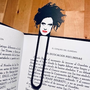 May include: Black metal bookmark with a stylized face and red lips, shaped like a paperclip. The bookmark is on a book with Spanish text. The face has dark hair.
