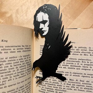 Puede incluir: Marcapáginas de papel negro con la silueta de un cuervo con rostro humano, inspirado en la película "The Crow". El marcapáginas está insertado en un libro con texto impreso. Un accesorio único para los amantes de los libros.