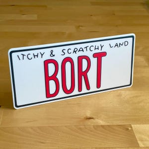以下が含まれることがあります： 「ITCHY & SCRATCHY LAND」の文字が上に、大きな赤い文字で「BORT」と書かれた白いナンバープレート。黒い縁取りがあり、木製の表面に置かれています。
