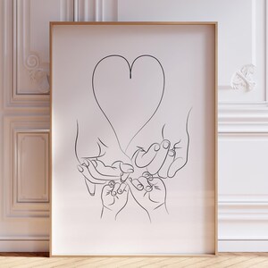 May include: A black and white line art print of hands forming a heart shape. The print is framed in a thin, gold frame and is hanging on a white wall.