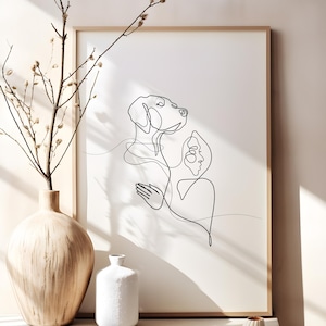 May include: A framed minimalist art print with a continuous line drawing of a dog and a person. The black artwork is set against a white background, in a light wood frame. A wooden vase with branches and two white ceramic vases are in the foreground.