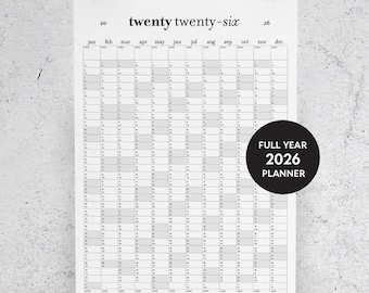 May include: A white 2026 full-year planner with a black circular sticker that reads "FULL YEAR 2026 PLANNER". The planner is held by two black binder clips. The text "twenty twenty-six" is at the top.