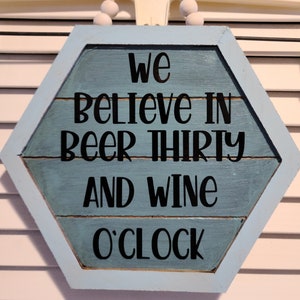 May include: A hexagonal wooden sign with a blue background and white trim. The sign reads "We believe in beer thirty and wine o'clock" in black lettering.