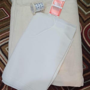 May include: A folded piece of off-white fabric layered beneath a piece of bright white fabric. A small spool of white thread with blue text and a package of Colonial brand needles with red and white packaging are placed on top of the fabrics.