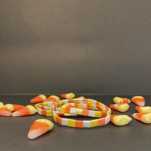 Puede incluir: Un conjunto de pulseras de maíz dulce con cuadrados blancos, amarillos y naranjas. Las pulseras están sobre una superficie gris con maíz dulce esparcido.