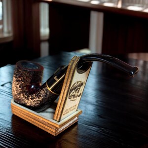 May include: A dark brown tobacco pipe with a textured, rustic bowl and a smooth black stem, resting in a light-colored wooden stand. The stand features a laser-engraved graphic of a skull wearing a hat and the text "STAND FOR SOMETHING."