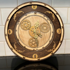 May include: A wooden clock with a dark brown outer ring and a light wood inner ring. The clock face has Roman numerals and a design of gears in the center. The clock hands are gold.