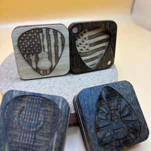 May include: Four wooden guitar pick holders with different designs. Two are American flag themed, one has a guitar design, and one has a sun design.