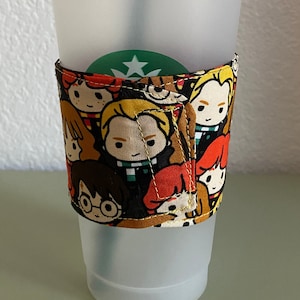 Può includere: Un bicchiere riutilizzabile in plastica trasparente con un logo verde e bianco e una fodera in tessuto con un motivo colorato di personaggi di Harry Potter. La fodera è nera con accenti rossi, gialli e arancioni.