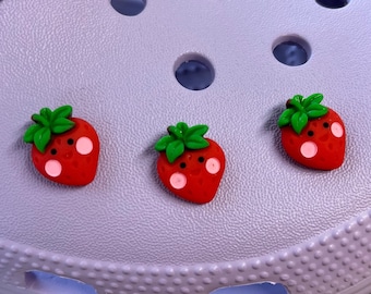 May include: Three red and green strawberry charms with white smiling faces. The charms are on a purple background.