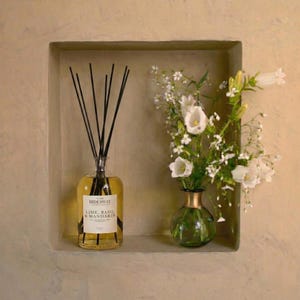 May include: A clear glass diffuser bottle with black reeds and a label that reads "HIDEAWAY LIME, BASIL & MANDARIN" sits in a recessed shelf. Beside it, a small green vase holds white flowers. The background is a neutral beige.