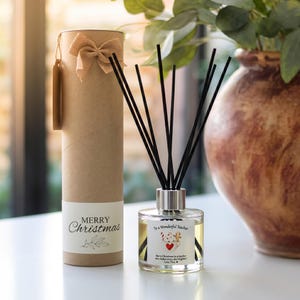 May include: A clear glass reed diffuser with black reeds and a label reading To a Wonderful Teacher, accompanied by a cylindrical kraft paper gift tube featuring a tan burlap bow and a label with the text Merry Christmas and leaf graphics.