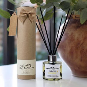 May include: A cylindrical brown cardboard gift tube with a burlap bow and a blank tag, paired with a glass reed diffuser containing yellow liquid and black reeds. The label reads: I'M A HAPPY-GO-LUCKY RAY OF FUCKING SUNSHINE. The tube says HAPPY Birthday.