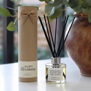 May include: A reed diffuser with black sticks in a clear glass bottle featuring a label that reads Mum said I had to get you a gift and Happy Birthday Claire, accompanied by a tall cylindrical brown cardboard gift tube with a burlap bow.