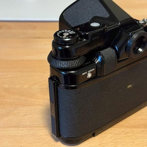 May include: A black vintage camera with a textured grip and various dials and buttons. The camera has a prominent viewfinder and a lens mount. The words "BATT. CHECK" are visible on the side. The camera is likely a film camera.