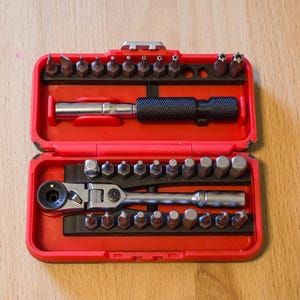 May include: A red toolbox containing a variety of metal screwdriver bits and a ratchet wrench. The set includes a black handled screwdriver and a silver ratchet wrench. The bits are arranged in rows within the open case.