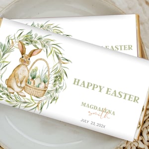 Könnte beinhalten: Ein weißer Schokoladenriegel-Wrapper mit einem grünen Aquarellkranz, der ein Kaninchen zeigt, das einen Korb mit Eiern hält. Der Text "Happy Easter" ist in grüner Schrift, gefolgt von "Magdalena Smith" in brauner Schrift und "July 23, 202X" in schwarzer Schrift.
