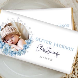 Op de afbeelding: Een witte chocolade reep met een blauw bloemenontwerp en een foto van een babyjongen. De tekst "Oliver Jackson Doop Juli 23, 202X" staat op de reep gedrukt.