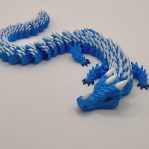 Könnte beinhalten: Eine blau-weiße, bewegliche Drachenfigur. Der Drache hat einen strukturierten Körper mit weißen Stacheln entlang des Rückens. Der Kopf des Drachen hat weiße Hörner und schwarze Krallen. Die Figur liegt auf einer weißen Oberfläche.