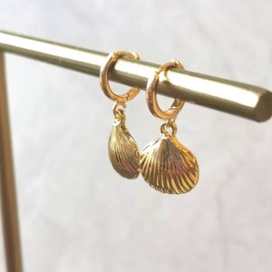 May include: Gold-plated hoop earrings featuring seashell pendants. The earrings are presented on a gold bar, set against a blurred backdrop. The seashell charms have detailed ridges and hang from the hoops.