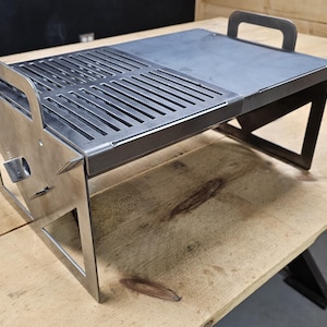 Può includere: Una piccola griglia portatile in metallo di forma rettangolare. Presenta una griglia e una superficie solida. La griglia ha una maniglia per il trasporto ed è posizionata su una superficie di legno. Dimensioni circa 40 cm x 30 cm.