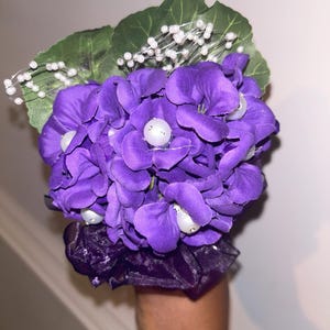 May include: A purple floral wrist corsage featuring a cluster of vibrant purple hydrangea-like flowers. The arrangement includes green leaves, white beaded accents, and a dark purple fabric wristband. A decorative accessory for special occasions.