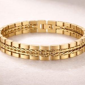 May include: A gold bracelet with a rectangular link design. The bracelet features a double-row chain with a secure clasp. The jewellery is displayed on a light-coloured surface.