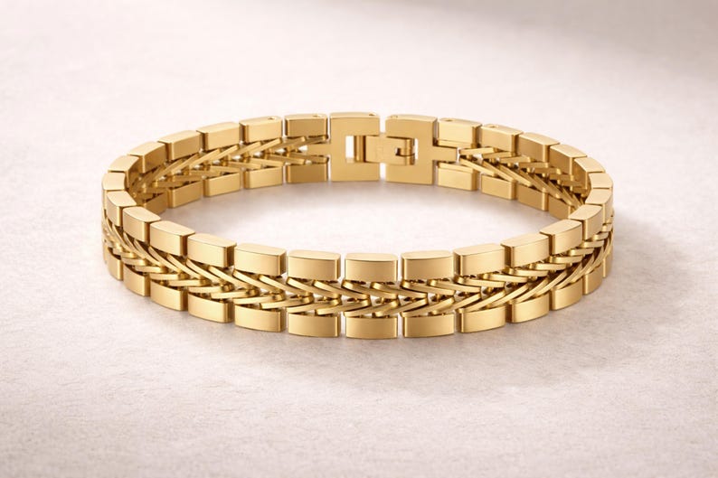 May include: A gold bracelet with a rectangular link design. The bracelet features a double-row chain with a secure clasp. The jewellery is displayed on a light-coloured surface.