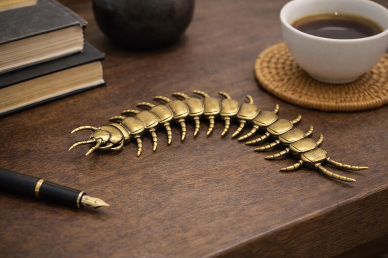 May include: A brass-colored centipede sculpture rests on a wooden surface, near a black pen and a cup of coffee. The detailed insect has multiple segments and legs, and is a decorative item.