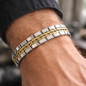 May include: A silver and gold bracelet featuring a woven gold central design and rectangular silver links. The bracelet is worn on a wrist, with a black shirt sleeve in the background. The jewellery is a stylish accessory.