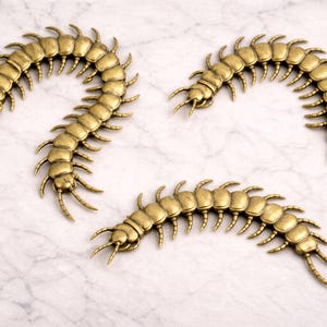 May include: Three antique gold-colored centipede decorative objects. Each centipede has a segmented body with numerous legs and antennae. The objects are arranged on a white and gray marble-patterned surface.
